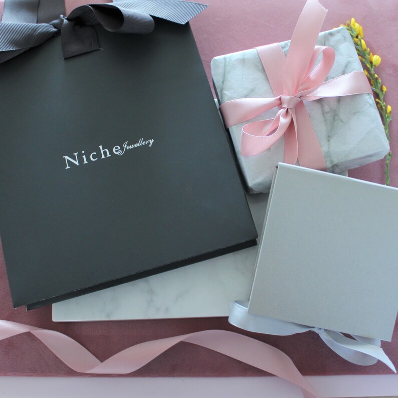 May include: Three gift boxes, one black with the text "Niche Jewellery", one white with pink marble pattern and a pink ribbon, and one gray with a white ribbon. All boxes are on a pink surface.