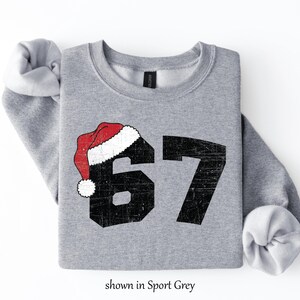 May include: A light grey sweatshirt with the number 67 in black, topped with a red and white Santa hat. The text "shown in Sport Grey" is at the bottom. The sweatshirt has rolled-up sleeves.
