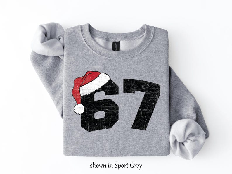 May include: A light grey sweatshirt with the number 67 in black, topped with a red and white Santa hat. The text "shown in Sport Grey" is at the bottom. The sweatshirt has rolled-up sleeves.