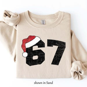 May include: Sand-colored sweatshirt with the number 67 in distressed black font, topped with a red and white Santa hat. The text "shown in Sand" is at the bottom. A cozy, casual top for the holiday season.