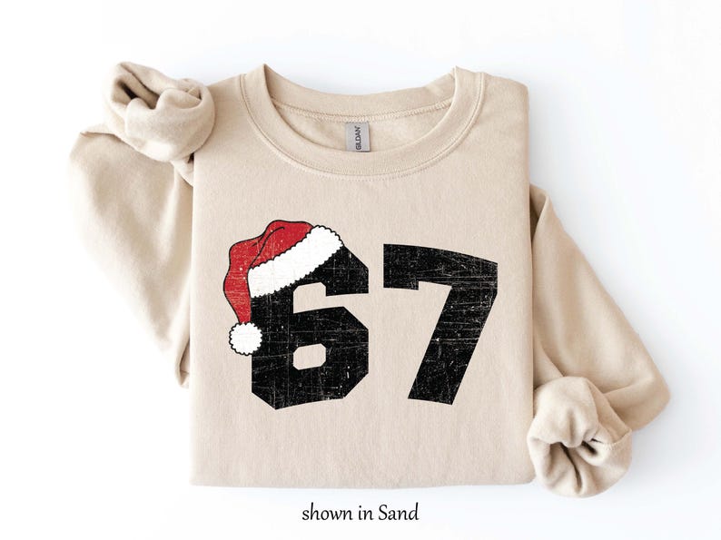 May include: Sand-colored sweatshirt with the number 67 in distressed black font, topped with a red and white Santa hat. The text "shown in Sand" is at the bottom. A cozy, casual top for the holiday season.