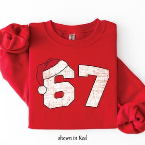 May include: Red sweatshirt with the number 67 in a distressed, off-white font, topped with a Santa hat. The text "shown in Red" is at the bottom. A festive, holiday-themed apparel item.
