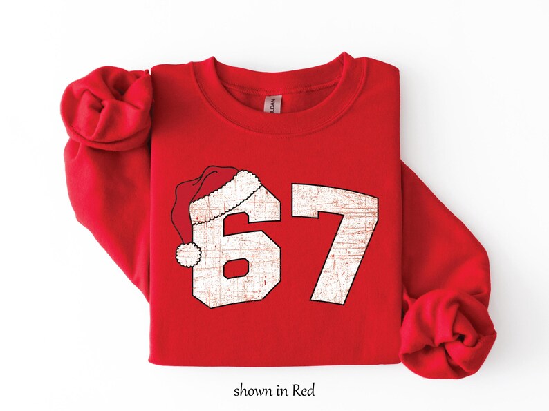 May include: Red sweatshirt with the number 67 in a distressed, off-white font, topped with a Santa hat. The text "shown in Red" is at the bottom. A festive, holiday-themed apparel item.