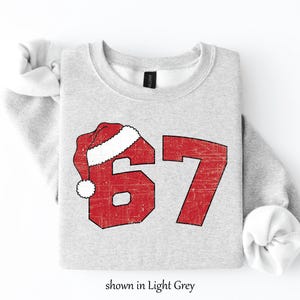 May include: Light grey sweatshirt with the number 67 in red, with a Santa hat over the 6. The text "shown in Light Grey" is at the bottom. The sweatshirt is a crew neck.