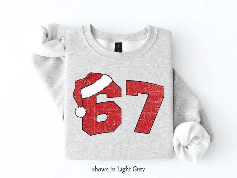 May include: Light grey sweatshirt with the number 67 in red, with a Santa hat over the 6. The text "shown in Light Grey" is at the bottom. The sweatshirt is a crew neck.