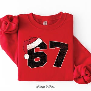 May include: A red sweatshirt with the number 67 in black, featuring a Santa hat over the number 6. The text "shown in Red" is at the bottom. The sweatshirt is a festive holiday apparel item.