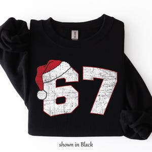 May include: Black sweatshirt with the number 67 in distressed white with red trim, and a Santa hat graphic. The text "shown in Black" is at the bottom of the image. A cozy, festive design.