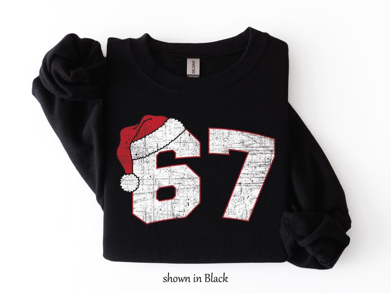May include: Black sweatshirt with the number 67 in distressed white with red trim, and a Santa hat graphic. The text "shown in Black" is at the bottom of the image. A cozy, festive design.