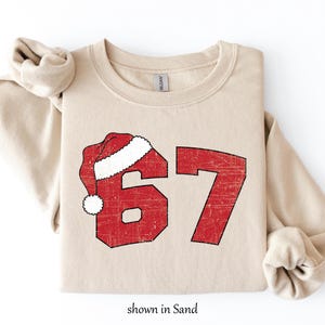 May include: A sand-colored sweatshirt with a red graphic of the number 67, topped with a Santa hat. The graphic has a distressed, vintage look. The text "shown in Sand" is at the bottom.