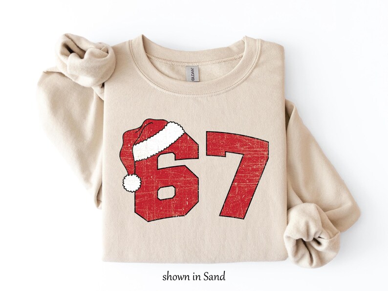 May include: A sand-colored sweatshirt with a red graphic of the number 67, topped with a Santa hat. The graphic has a distressed, vintage look. The text "shown in Sand" is at the bottom.