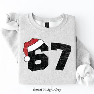 May include: Light grey sweatshirt with the number 67 in distressed black font, topped with a red and white Santa hat. The text "shown in Light Grey" is at the bottom of the image. A festive holiday-themed apparel item.