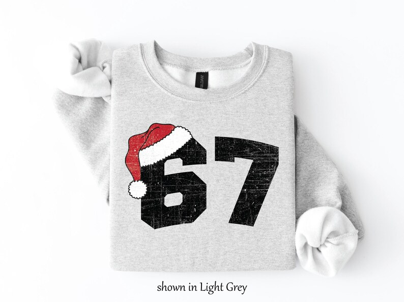 May include: Light grey sweatshirt with the number 67 in distressed black font, topped with a red and white Santa hat. The text "shown in Light Grey" is at the bottom of the image. A festive holiday-themed apparel item.