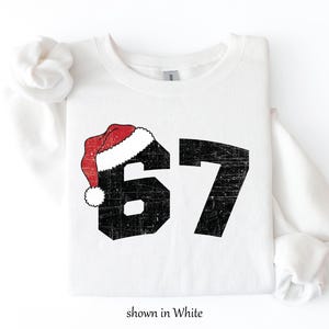 May include: White sweatshirt with the number 67 in black, distressed font. A red and white Santa hat is on top of the number 6. The text "shown in White" is at the bottom.