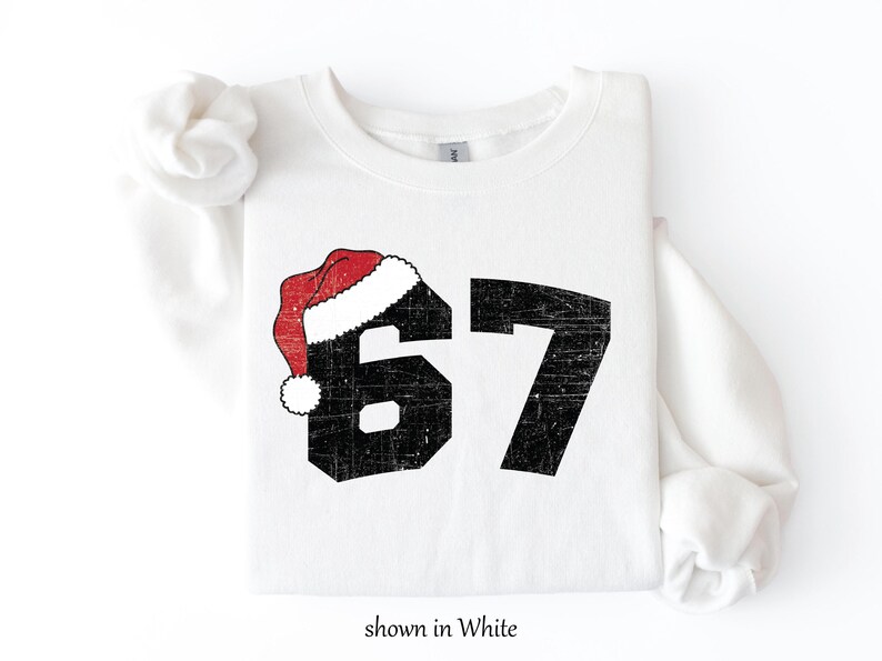 May include: White sweatshirt with the number 67 in black, distressed font. A red and white Santa hat is on top of the number 6. The text "shown in White" is at the bottom.
