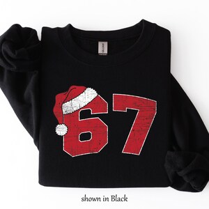 May include: Black sweatshirt with a distressed red number 67 design, topped with a Santa hat. The text "shown in Black" is at the bottom. The sweatshirt is folded on a white surface.
