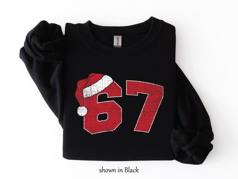 May include: Black sweatshirt with a distressed red number 67 design, topped with a Santa hat. The text "shown in Black" is at the bottom. The sweatshirt is folded on a white surface.