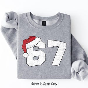 May include: A light gray sweatshirt with the number 67 in white, topped with a red and white Santa hat. The text "shown in Sport Grey" is at the bottom. The sweatshirt has rolled-up sleeves.