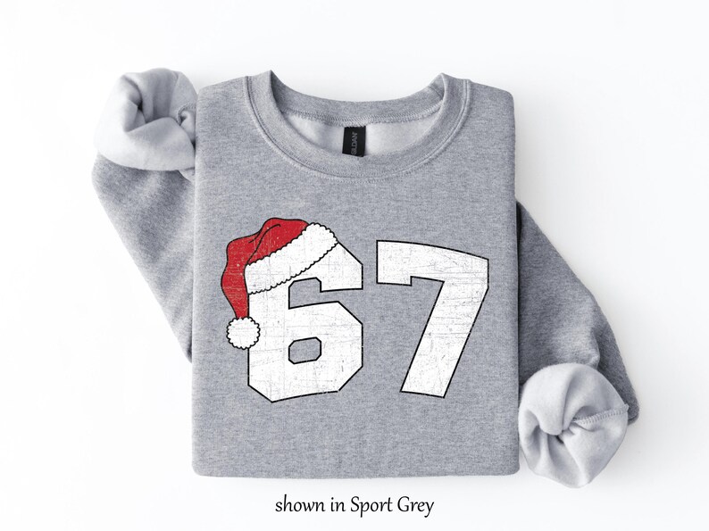 May include: A light gray sweatshirt with the number 67 in white, topped with a red and white Santa hat. The text "shown in Sport Grey" is at the bottom. The sweatshirt has rolled-up sleeves.