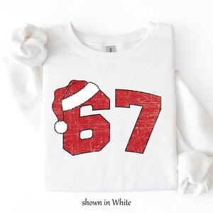 May include: White sweatshirt with a distressed red graphic of the number 67, topped with a Santa hat. The text "shown in White" is at the bottom. A festive, holiday-themed apparel item.
