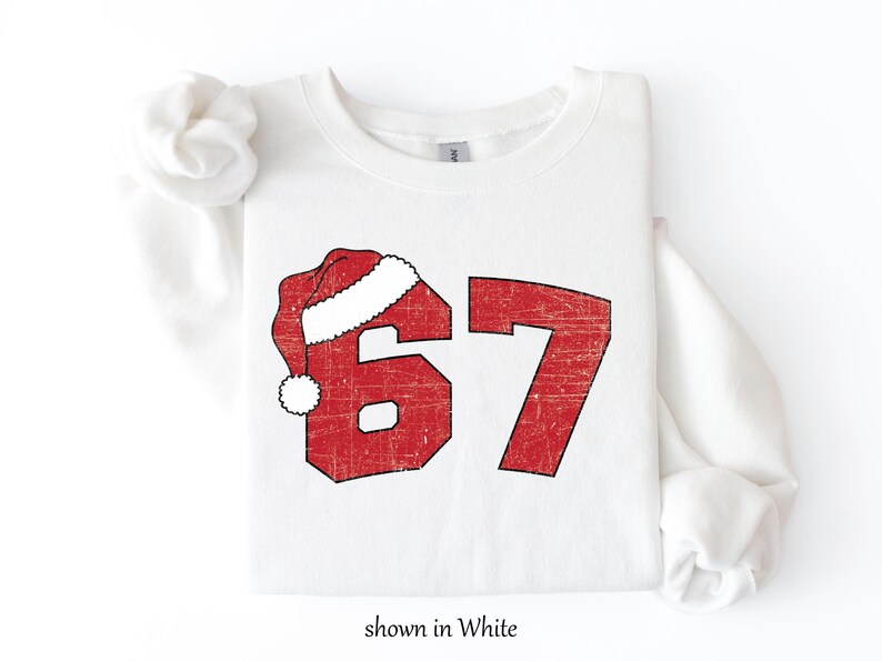 May include: White sweatshirt with a distressed red graphic of the number 67, topped with a Santa hat. The text "shown in White" is at the bottom. A festive, holiday-themed apparel item.