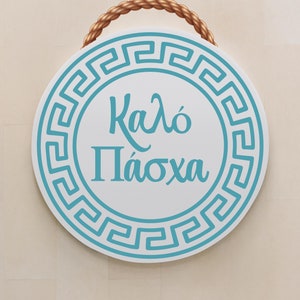 May include: A white round sign with a blue Greek key border and the text "Kaló Páсχα" in blue cursive lettering. The sign is hanging from a brown rope.