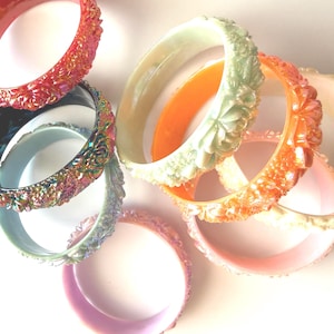 May include: A collection of colorful bangles in various shades, including red, iridescent, green, orange, and pink. The bangles have a textured, floral design. The jewelry is stacked and arranged on a white surface.