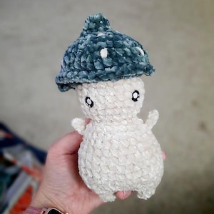 Puede incluir: Un juguete de peluche tejido a crochet con forma de hongo. El hongo tiene un cuerpo blanco y un gorro azul verdoso con lunares blancos. El juguete tiene ojos negros y un cuerpo pequeño y redondeado.