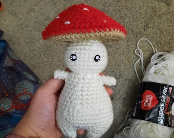 May include: White crocheted mushroom amigurumi with red and white spotted cap. The mushroom is being held by a hand wearing a pink watch.