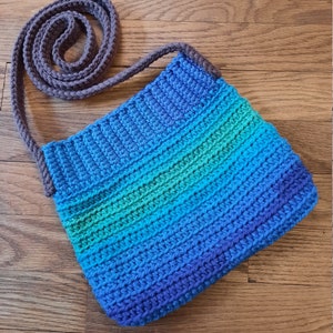 May include: A crocheted shoulder bag with a blue, green, and purple ombre pattern. The bag has a brown crocheted strap.