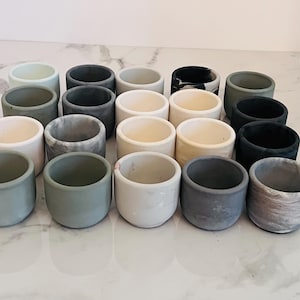 Peut inclure: Une collection de 18 petits pots ronds en béton de différentes couleurs, dont le blanc, le gris, le vert et le noir. Certains pots ont un design marbré.