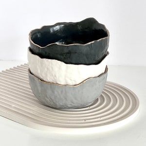May include: Three ceramic bowls stacked on top of each other. The top bowl is black, the middle bowl is white, and the bottom bowl is grey. All three bowls have a gold rim.