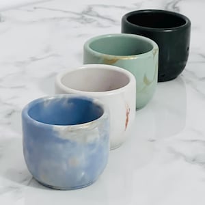 May include: Four small ceramic cups in a row, each with a unique colour and design. The cups are blue, white, mint green, and dark green. The blue cup has a marbled effect, the white cup has a red streak, and the mint green cup has gold accents.