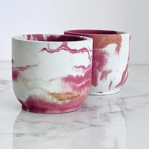 May include: Two cylindrical planters with a marbled design. The planters are white with swirls of pink, gold, and peach. The interior and rim of the planters are a solid pink color. The planters are sitting on a reflective surface.