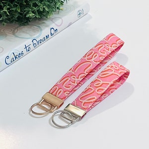 Peut inclure: Deux porte-clés roses avec des anneaux de clés dorés et argentés. Les porte-clés sont en tissu avec un motif en spirale rose et blanc.