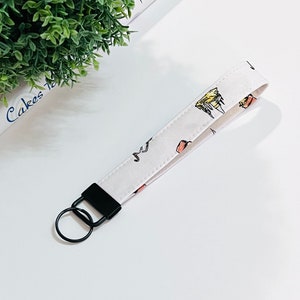 May include: White fabric keychain with a black metal ring and a black clip. The fabric has a pattern of small, colourful illustrations.