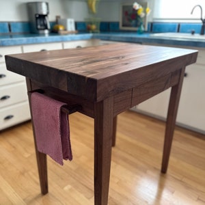 May include: A dark brown wooden kitchen island with a butcher block countertop and a towel rack on the side. The island has a drawer and is standing on a hardwood floor.