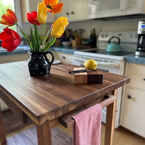 May include: A wooden kitchen island with a walnut finish. A black and white polka-dotted pitcher holds a bouquet of red, orange, and yellow tulips. A small cutting board with a lemon and knife sits on top. A red and white checkered towel hangs below.
