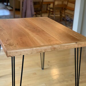 May include: A light wood table with a natural finish and a live edge top. The table has four black hairpin legs.