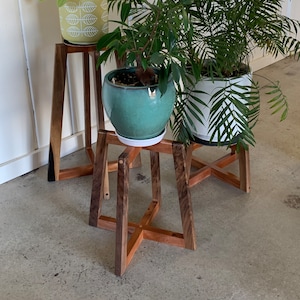 May include: Three wooden plant stands with different sized plants in pots. The stands are made of dark wood and have a simple, modern design. The plants are a green leafy plant, a yellow plant with white leaves, and a white plant with green leaves.