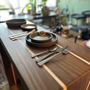 May include: A wooden table with a light wood inlay, set with two dark gray ceramic plates, a small bowl, and silver cutlery.