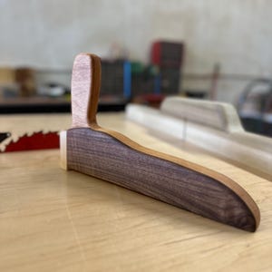 May include: A handcrafted wooden bread lame with a curved blade holder and a handle. The blade holder is made of dark walnut, and the handle is made of a lighter wood. The bread lame is designed for scoring bread dough.