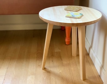 May include: A small, round wooden end table with four legs. The table is made of light wood and has a natural finish. There are two coasters on the table, one with a yellow and orange pattern and the other with a lemon pattern.