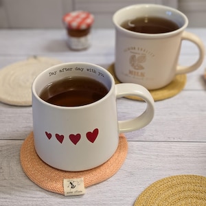 Può includere: Due tazze in ceramica bianca con manici, una con il testo "Day after day with you" e tre cuori rossi, l'altra con il testo "Our Family Milk". Entrambe le tazze sono riempite di liquido marrone e sono appoggiate su sottobicchieri rotondi intrecciati.