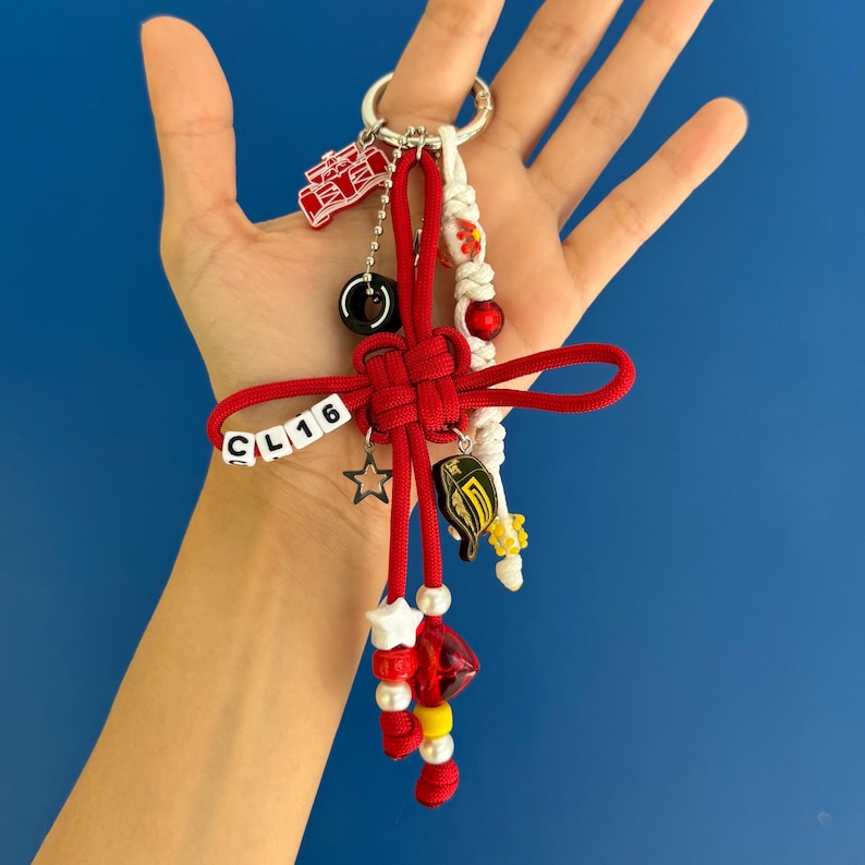 May include: A red and white keychain with various charms, including a star, a black circle, and a small red car. The keychain also features the letters "CL16" on white beads and a red knot design.