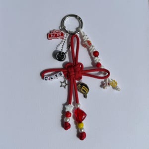 May include: A red and white keychain with various charms, including a race car, a star, and a baseball glove. The keychain features a knotted design with the letters "CL16" on small white beads. Additional red and white beads and a silver keyring complete the design.