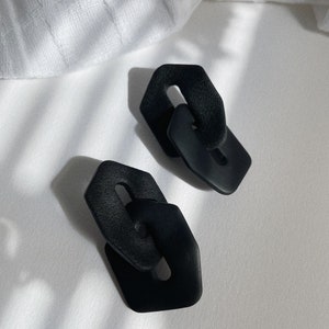 May include: A pair of black geometric earrings with a linked design. The earrings are made of a textured material and have a matte finish.