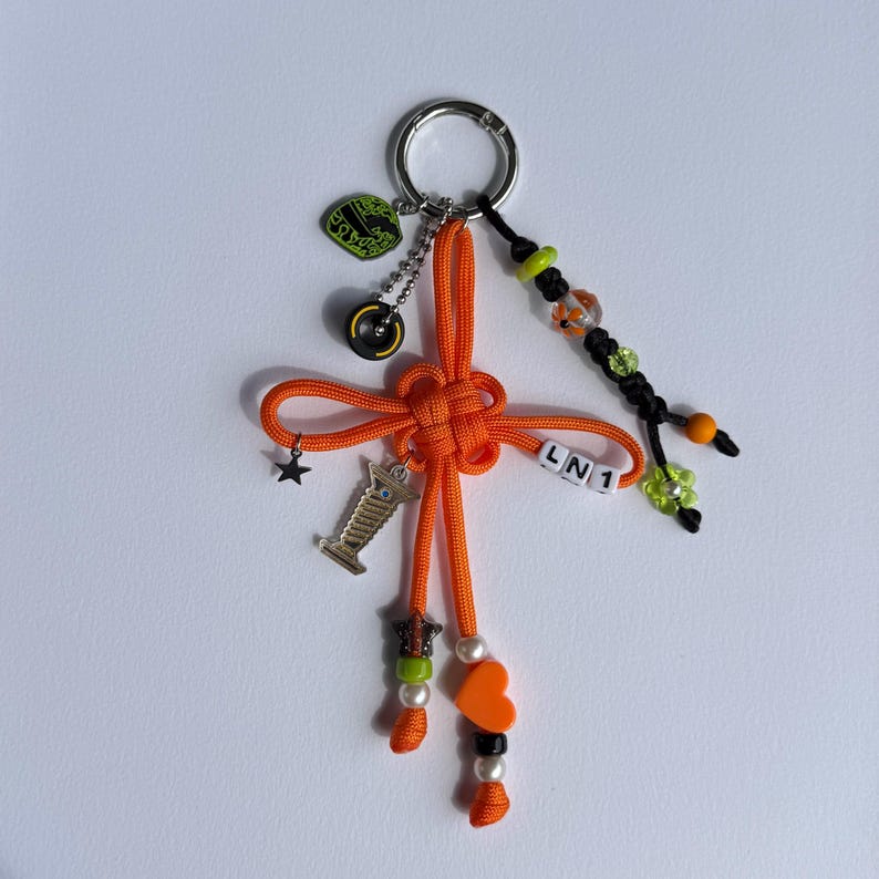 May include: Orange keychain with a knotted design, adorned with charms including a star, a race car, and a heart. Additional details include beads, a small crown, and the letters "LN1". The keychain has a silver ring for attaching keys.