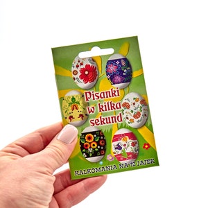 May include: A package of Easter egg decals held by a hand. The green card features six decorated egg designs with floral patterns in red, blue, yellow, and orange. Text on the card reads "Pisanki w kilka sekund" and "Kalkomania na 12 jajek".