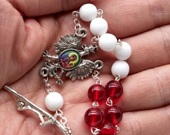 May include: A white and red rosary with a silver charm featuring a Polish eagle and a religious image. The beads are made of white and red glass.