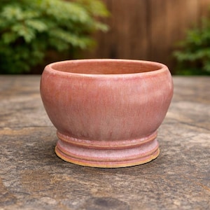 May include: A small, round ceramic bowl with a smooth, glossy finish in shades of dusty rose and soft pink. The bowl features a wide, curved body that tapers down to a distinct, raised circular base with a visible light tan rim at the bottom edge.